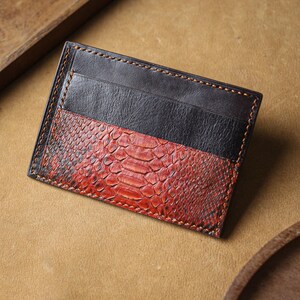 Puede incluir: Un tarjetero rectangular con un dise&ntilde;o de dos tonos. La secci&oacute;n superior es de cuero negro liso, mientras que la secci&oacute;n inferior muestra un patr&oacute;n texturizado de piel de serpiente roja y negra. La pieza est&aacute; rematada con costuras naranjas visibles.