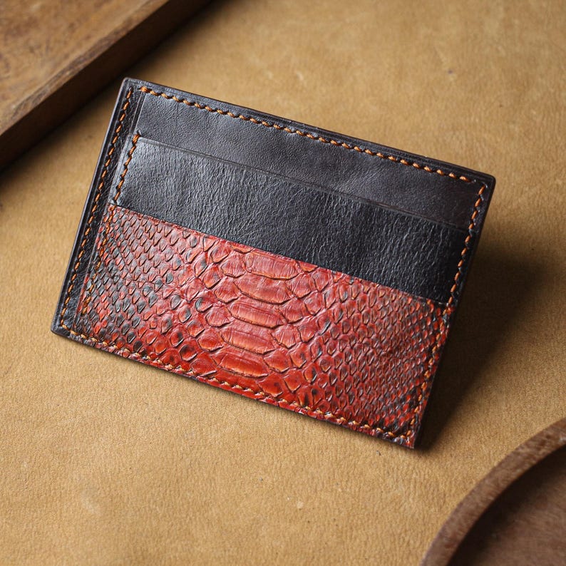 Puede incluir: Un tarjetero rectangular con un dise&ntilde;o de dos tonos. La secci&oacute;n superior es de cuero negro liso, mientras que la secci&oacute;n inferior muestra un patr&oacute;n texturizado de piel de serpiente roja y negra. La pieza est&aacute; rematada con costuras naranjas visibles.