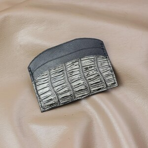 May include: A slim card holder featuring a dark grey cross-hatched texture on the upper section and a metallic silver-toned crocodile skin pattern on the lower section. The accessory has a curved top edge and visible stitching along the perimeter.