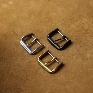 Pode incluir: Três fivelas de relógio de metal em prata, preto e dourado. As fivelas são retangulares com uma única barra para a pulseira do relógio. O fundo é uma superfície castanha texturizada.