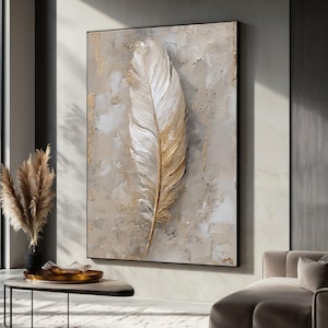 Op de afbeelding: Een groot, ingelijst canvas kunstwerk met een getextureerde, driedimensionale veer. De veer is voornamelijk wit met gouden accenten, tegen een neutrale, getextureerde achtergrond. Het kunstwerk is omlijst door een zwarte rand.