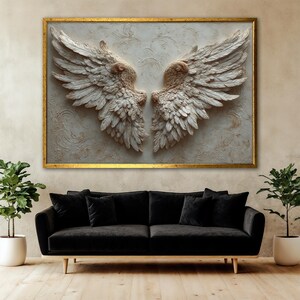 May include: A large framed artwork featuring a pair of sculpted angel wings in shades of white and beige. The wings are detailed with individual feathers and set against a textured background. The frame is gold, and the artwork hangs above a black sofa.