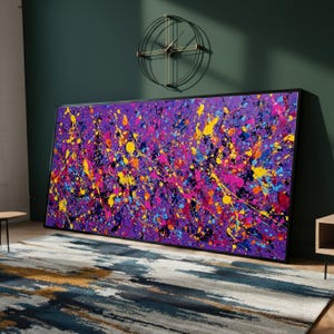 May include: Large abstract painting with a black frame. The artwork features a vibrant display of colorful paint splatters in shades of purple, pink, yellow, and blue. The painting leans against a dark green wall, with a modern clock and rug in the background.
