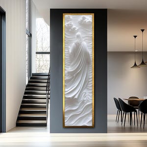 K&ouml;nnte beinhalten: Wei&szlig;e 3D-Wandskulptur einer Person in einem flie&szlig;enden Gewand, in Gold gerahmt mit wei&szlig;em Hintergrund. Die Skulptur ist an einer grauen Wand montiert.