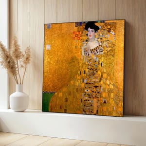May include: A framed print of a Gustav Klimt painting. The artwork features a woman in a gold and patterned dress, set against a gold background. The painting is displayed on a white ledge with a vase and dried plants.