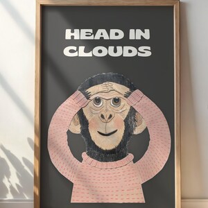 Op de afbeelding: Een ingelijste kunstdruk met een cartoon aap met zijn handen voor zijn ogen. De aap draagt een roze trui en de woorden "HEAD IN CLOUDS" staan er in het wit boven. De lijst is van hout.