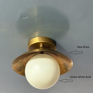 May include: A ceiling-mounted light fixture with a raw brass finish and a globe white bulb. The fixture has a circular base and a flared shade, casting a warm glow. The fixture is mounted on a light blue ceiling.