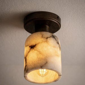 Puede incluir: Una l&aacute;mpara de techo con una pantalla cil&iacute;ndrica con aspecto de m&aacute;rmol. La pantalla es de color blanco crema con vetas marrones, y la l&aacute;mpara tiene una base de color bronce oscuro. La bombilla es visible dentro de la pantalla.