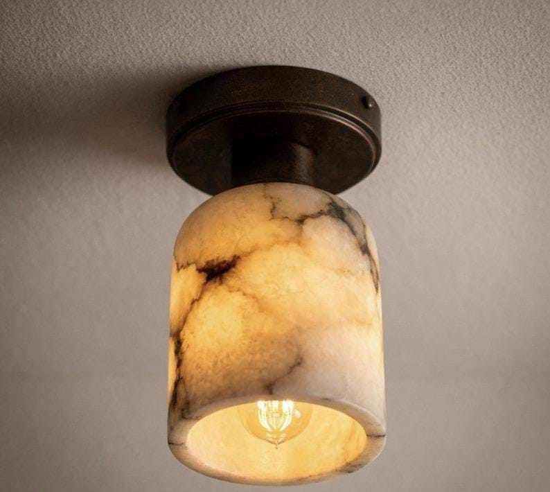 Puede incluir: Una l&aacute;mpara de techo con una pantalla cil&iacute;ndrica con aspecto de m&aacute;rmol. La pantalla es de color blanco crema con vetas marrones, y la l&aacute;mpara tiene una base de color bronce oscuro. La bombilla es visible dentro de la pantalla.