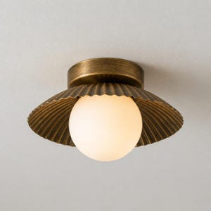 May include: A brass-toned ceiling light fixture with a seashell-inspired design. The fixture features a round, frosted glass globe light, surrounded by a scalloped, textured metal shade. The light is mounted on a circular base.