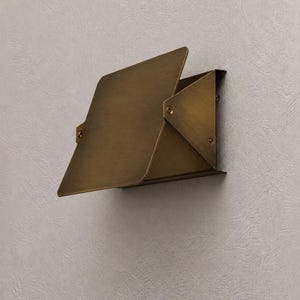 May include: A brass-colored wall-mounted light fixture with a geometric design. The fixture has a rectangular base and a hinged, angled shade. The shade is made of folded metal and is attached to the base with rivets.