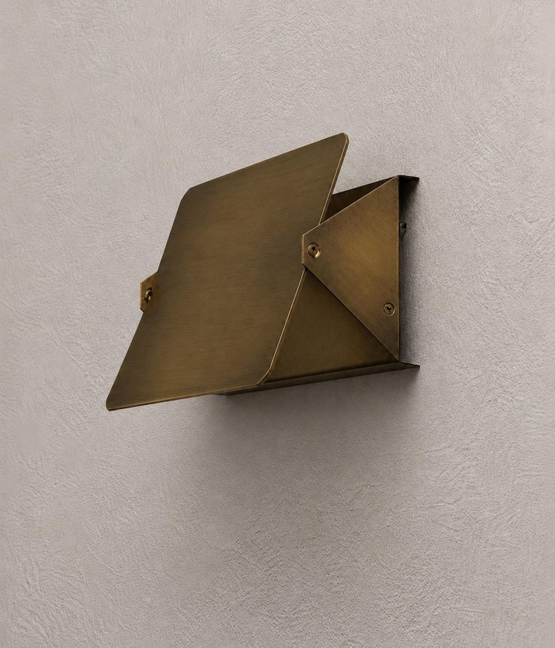 May include: A brass-colored wall-mounted light fixture with a geometric design. The fixture has a rectangular base and a hinged, angled shade. The shade is made of folded metal and is attached to the base with rivets.