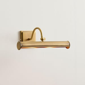 Peut inclure: Une applique murale pour tableau avec une finition en laiton brossé. La lampe comprend une plaque arrière rectangulaire, un bras incurvé réglable et un abat-jour cylindrique horizontal qui dirige la lumière vers le bas pour éclairer les œuvres.