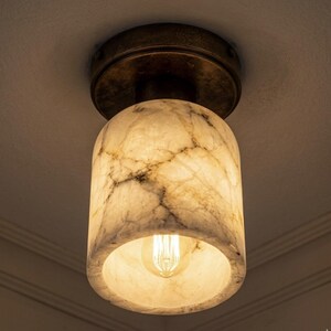 Puede incluir: Una l&aacute;mpara de techo con una pantalla cil&iacute;ndrica con efecto m&aacute;rmol. La pantalla es blanquecina con vetas grises, y la base es de color bronce. Una bombilla c&aacute;lida es visible dentro de la pantalla, iluminando el espacio.