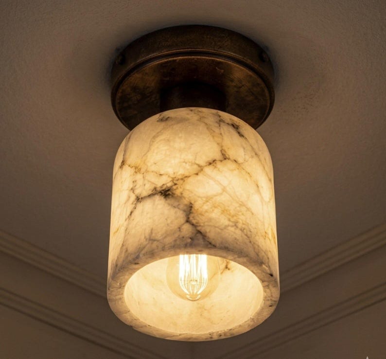Puede incluir: Una l&aacute;mpara de techo con una pantalla cil&iacute;ndrica con efecto m&aacute;rmol. La pantalla es blanquecina con vetas grises, y la base es de color bronce. Una bombilla c&aacute;lida es visible dentro de la pantalla, iluminando el espacio.