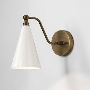 May include: A modern wall sconce with a white, cone-shaped shade and a brass-colored arm and circular base. The sconce is mounted on a white wall, providing a clean and minimalist aesthetic. The light fixture is designed for indoor use.
