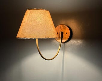 May include: A wall-mounted lamp featuring a tan fabric lampshade and a brass-toned metal frame. The lamp has a circular backplate and a curved arm supporting the shade. The shade is cone-shaped with a textured finish. The lamp is designed for wall mounting.