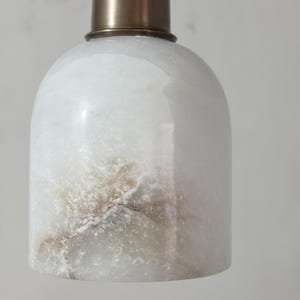 Puede incluir: Una l&aacute;mpara colgante de &oacute;nix blanco transl&uacute;cido con una tapa de metal color lat&oacute;n. La piedra tiene vetas naturales marrones y grises. La l&aacute;mpara tiene una forma redondeada, similar a una campana.