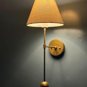 Puede incluir: Aplique de pared de latón con pantalla de tela beige. El aplique tiene un soporte de pared redondo, un brazo vertical largo y un peso esférico. La pantalla cónica emite una luz cálida.