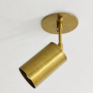May include: A brass-colored, cylindrical ceiling spotlight fixture. The light has a round base and a pivoting arm, allowing for directional lighting. The fixture is mounted on a white ceiling.