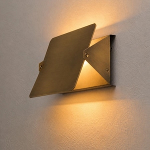 May include: A modern, geometric wall sconce with a brass finish. The fixture has a rectangular base and a pivoting, angled shade that directs light downwards. The sconce is mounted on a textured, off-white wall, creating a warm, inviting ambiance.