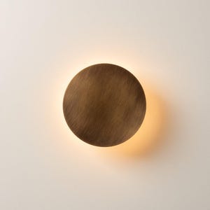 Puede incluir: Un aplique de pared circular con acabado en bronce cepillado. La placa metálica en forma de disco está centrada y emite un brillo cálido y suave desde detrás de sus bordes, creando un efecto de halo sobre la superficie de la pared.