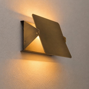 May include: A modern, geometric wall sconce with a brass finish. The fixture has a square shape with a hinged, angled panel that partially covers the light source, creating a warm, inviting glow. The sconce is mounted on a textured, light-colored wall.