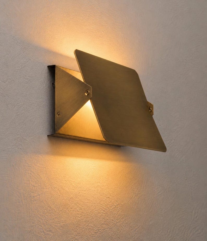 May include: A modern, geometric wall sconce with a brass finish. The fixture has a square shape with a hinged, angled panel that partially covers the light source, creating a warm, inviting glow. The sconce is mounted on a textured, light-colored wall.