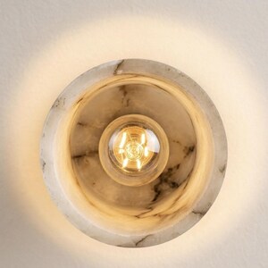 Puede incluir: Aplique de pared redondo de m&aacute;rmol con una bombilla central. La l&aacute;mpara tiene un dise&ntilde;o circular con una bombilla en el centro. El m&aacute;rmol presenta un patr&oacute;n veteado natural en tonos blancos y grises. La bombilla emite una luz c&aacute;lida y dorada.