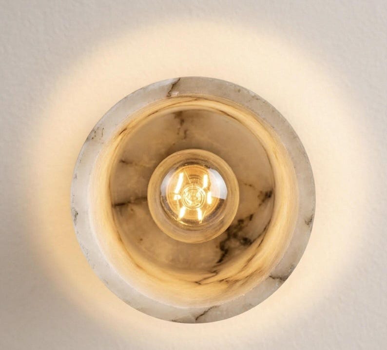 Puede incluir: Aplique de pared redondo de m&aacute;rmol con una bombilla central. La l&aacute;mpara tiene un dise&ntilde;o circular con una bombilla en el centro. El m&aacute;rmol presenta un patr&oacute;n veteado natural en tonos blancos y grises. La bombilla emite una luz c&aacute;lida y dorada.
