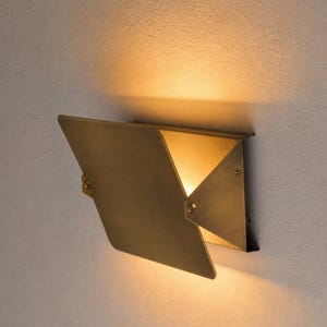 May include: A modern, geometric wall sconce with a brass finish. The fixture has a unique, origami-like design with a hinged, triangular shade that directs warm light downwards. The sconce is mounted on a light-colored wall.
