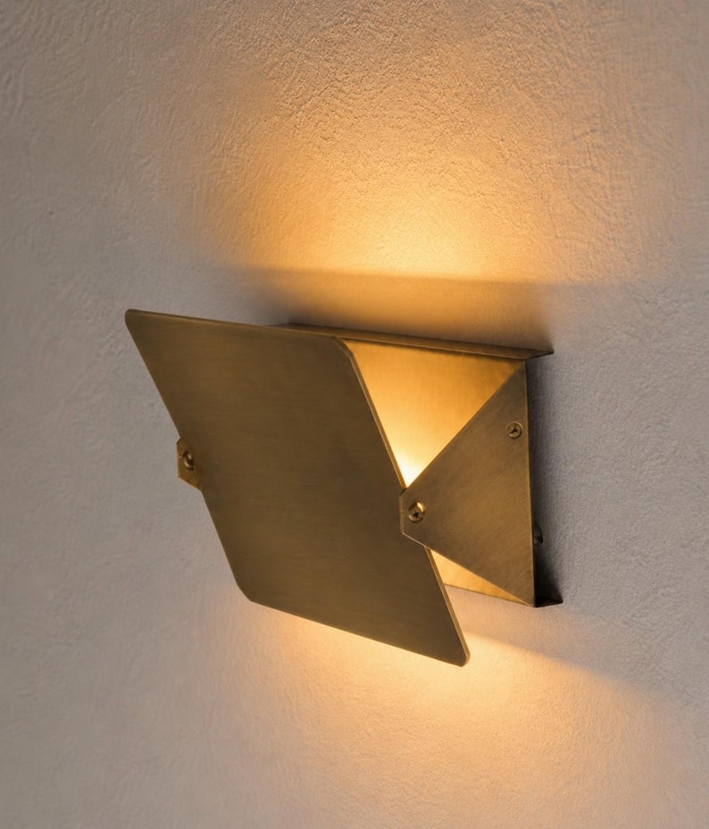 May include: A modern, geometric wall sconce with a brass finish. The fixture has a unique, origami-like design with a hinged, triangular shade that directs warm light downwards. The sconce is mounted on a light-colored wall.