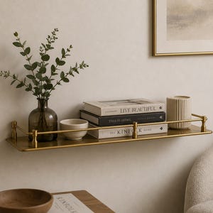 Op de afbeelding: Een rechthoekige wandplank van goudkleurig metaal met een beschermende voorstang. De plank bevat een donkere glazen vaas met groen blad, een kleine witte kom, een stapel van drie boeken met de titels Live Beautiful, This Is Home en The Kinfolk Home, en een geribbelde witte kaars.