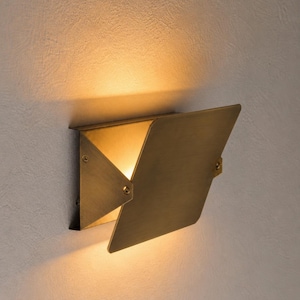 May include: A brass-colored wall sconce with a geometric design. The sconce has a hinged, angled cover that opens to reveal a warm light. The fixture is mounted on a textured, light-colored wall.