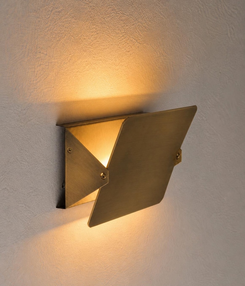 May include: A brass-colored wall sconce with a geometric design. The sconce has a hinged, angled cover that opens to reveal a warm light. The fixture is mounted on a textured, light-colored wall.