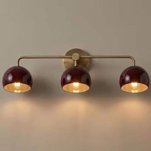 May include: A three-light wall fitting featuring a horizontal brass arm and a circular wall mount. Three glossy, deep burgundy, dome-shaped shades are attached to the arm, each housing a glowing light bulb with a reflective gold interior finish.