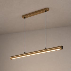 May include: A linear pendant light fixture featuring a long, cylindrical gold-toned metal tube suspended by two thin black cords from a matching rectangular gold-toned ceiling canopy. The light emits a warm glow from the underside of the tube.
