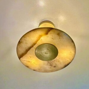 May include: A circular wall-mounted light fixture featuring a translucent, cream-colored stone shade with natural brown veining. The center of the disc contains a smaller, solid circular element in a muted olive green tone, emitting a warm glow.