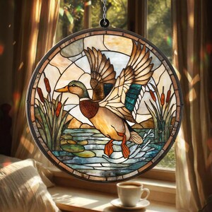 Puede incluir: Una vidriera redonda que representa un pato en vuelo sobre agua, juncos y nenúfares. El pato tiene plumas marrones, turquesas y blancas. La ventana está enmarcada en metal marrón oscuro y cuelga de una cadena.
