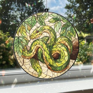 Könnte beinhalten: Runde Glasmalerei mit einer grünen Schlange, die sich um einen braunen Ast windet, umgeben von grünen Blättern. Die Schlange hat rote Augen und ist vor einem hellen Hintergrund platziert. Das Kunstwerk ist in einem schwarzen Metallring gerahmt.