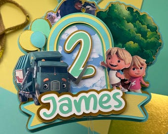 May include: A colorful cake topper featuring a green garbage truck, cartoon characters, and the number 2. The name "James" is displayed on a banner. The topper has a gold trim and is set against a yellow and teal background.