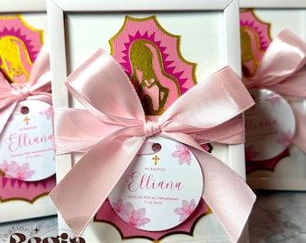 May include: A framed gift box with a pink ribbon and tag. The tag reads "Elliana" and "Mi Bautizo" with a cross and floral accents. The box features a pink and gold religious design.