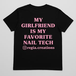 May include: Black t-shirt with the text "MY GIRLFRIEND IS MY FAVORITE NAIL TECH" in pink, along with the Instagram handle "@regia.creations". The shirt is a classic crew neck style.