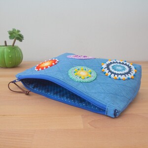 May include: A blue zippered pouch with a dotted lining. The pouch is decorated with four colourful crocheted circles.