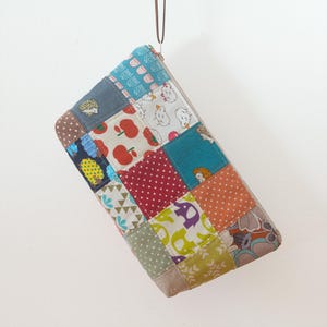 May include: A patchwork wallet with a variety of fabric squares in different colours and patterns. The wallet features a brown wrist strap and a zip closure. The fabrics include polka dots, apples, and animal prints.