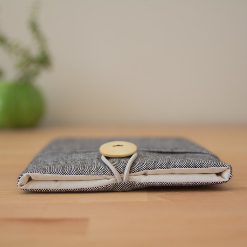 May include: A gray linen laptop sleeve with a wooden button closure. The sleeve is folded closed and sitting on a wooden surface.