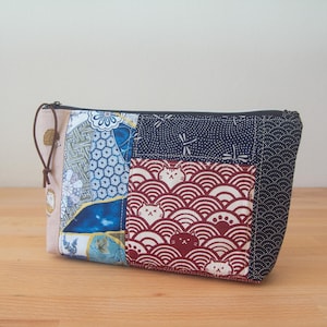 Op de afbeelding: Een patchwork etui met verschillende patronen. De etui heeft een bruine leren trekker, een marineblauwe rits en een zak met een rood en wit kattenpatroon. De stoffen omvatten bloemen-, geometrische en golfmotieven.
