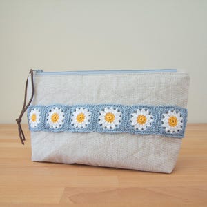 May include: A light beige zippered pouch with a quilted design and a crocheted trim of white and yellow daisy flowers. The trim is a light blue color.