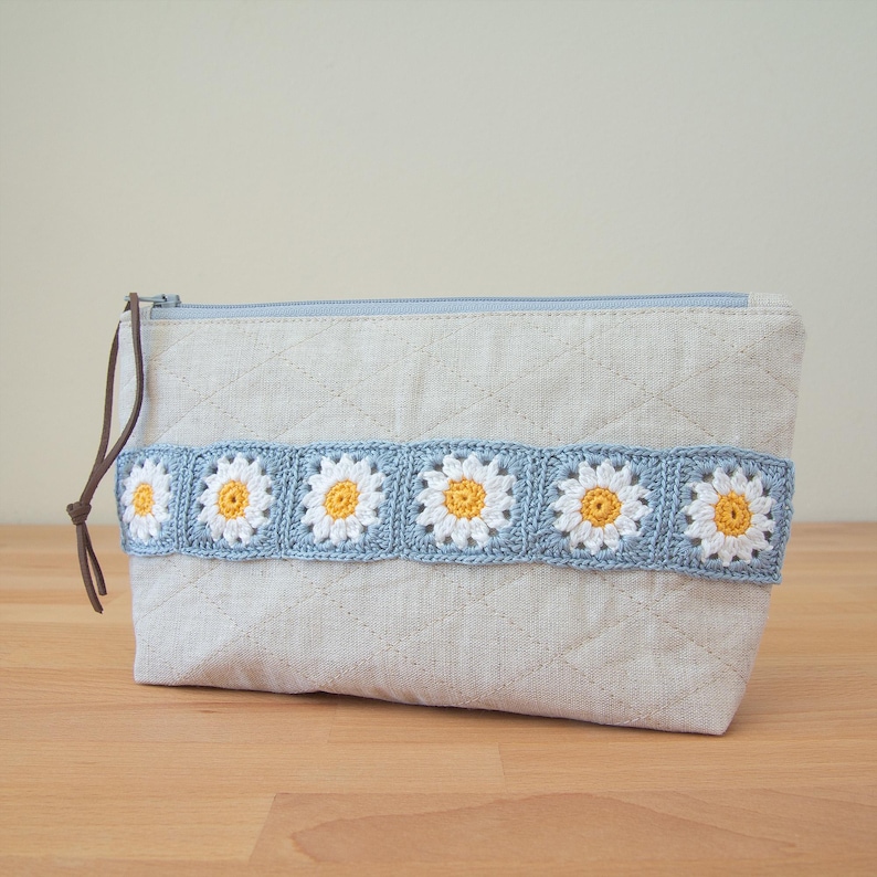 May include: A light beige zippered pouch with a quilted design and a crocheted trim of white and yellow daisy flowers. The trim is a light blue color.