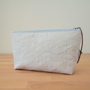 May include: A light blue zippered pouch with a quilted linen exterior. The pouch is made of a natural fabric and has a brown leather pull tab.